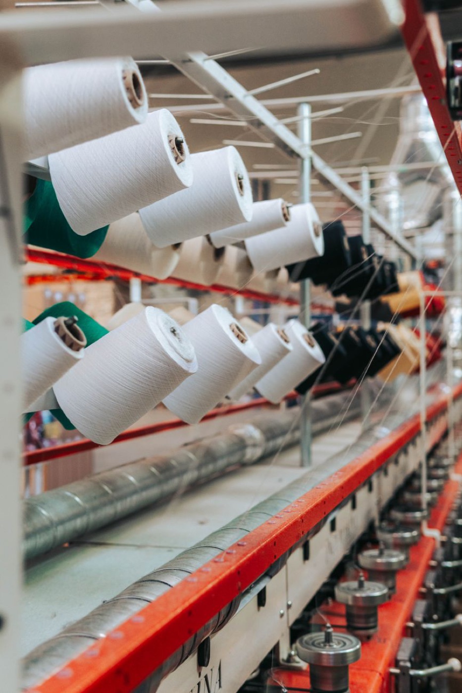 Rows of white yarn cones mounted on a red and cream twisting frame in a textile mill, fine threads running down to spindles below