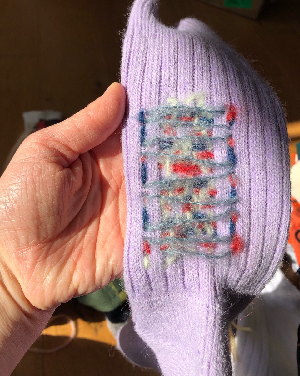 A lilac ribbed sock held to the window, its heel rewoven in red, cream and blue by London textile artist Celia Pym