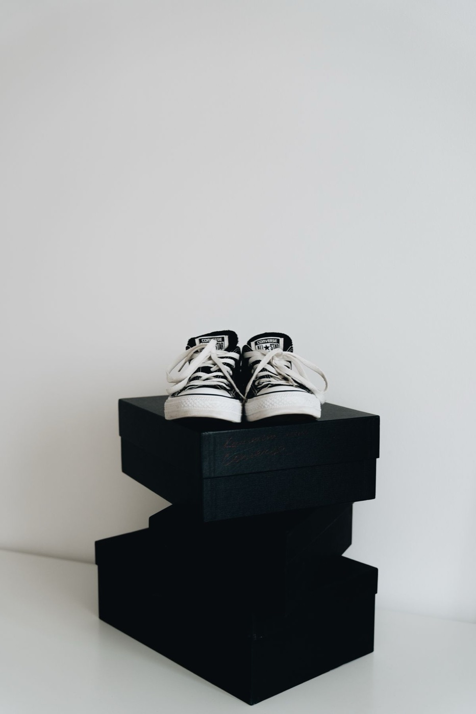 A pair of black low-top sneakers with white laces sitting on a stack of black shoe boxes against a white wall