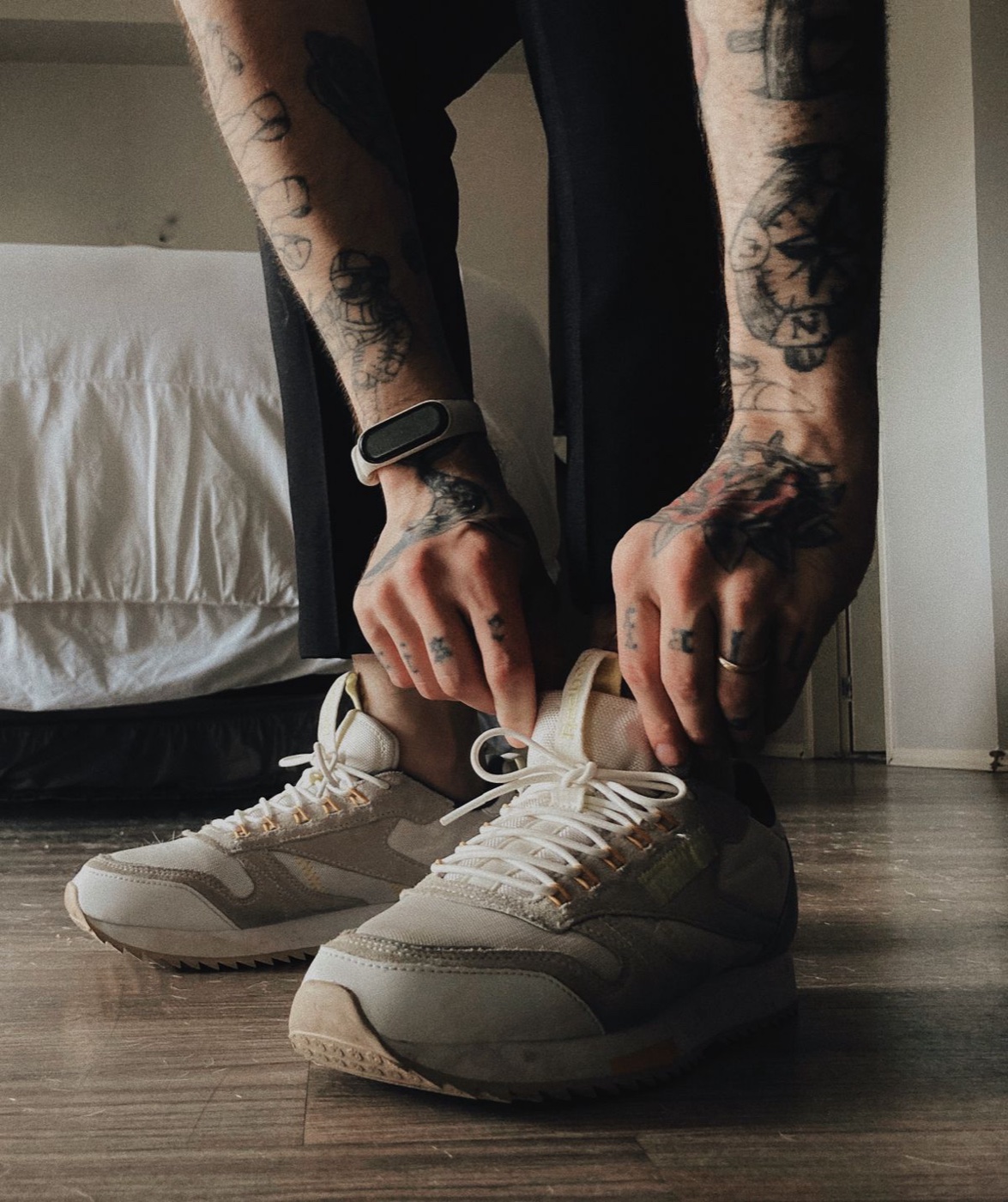 A person bending down to tie the laces of beige Reebok sneakers on a wooden floor beside a bed