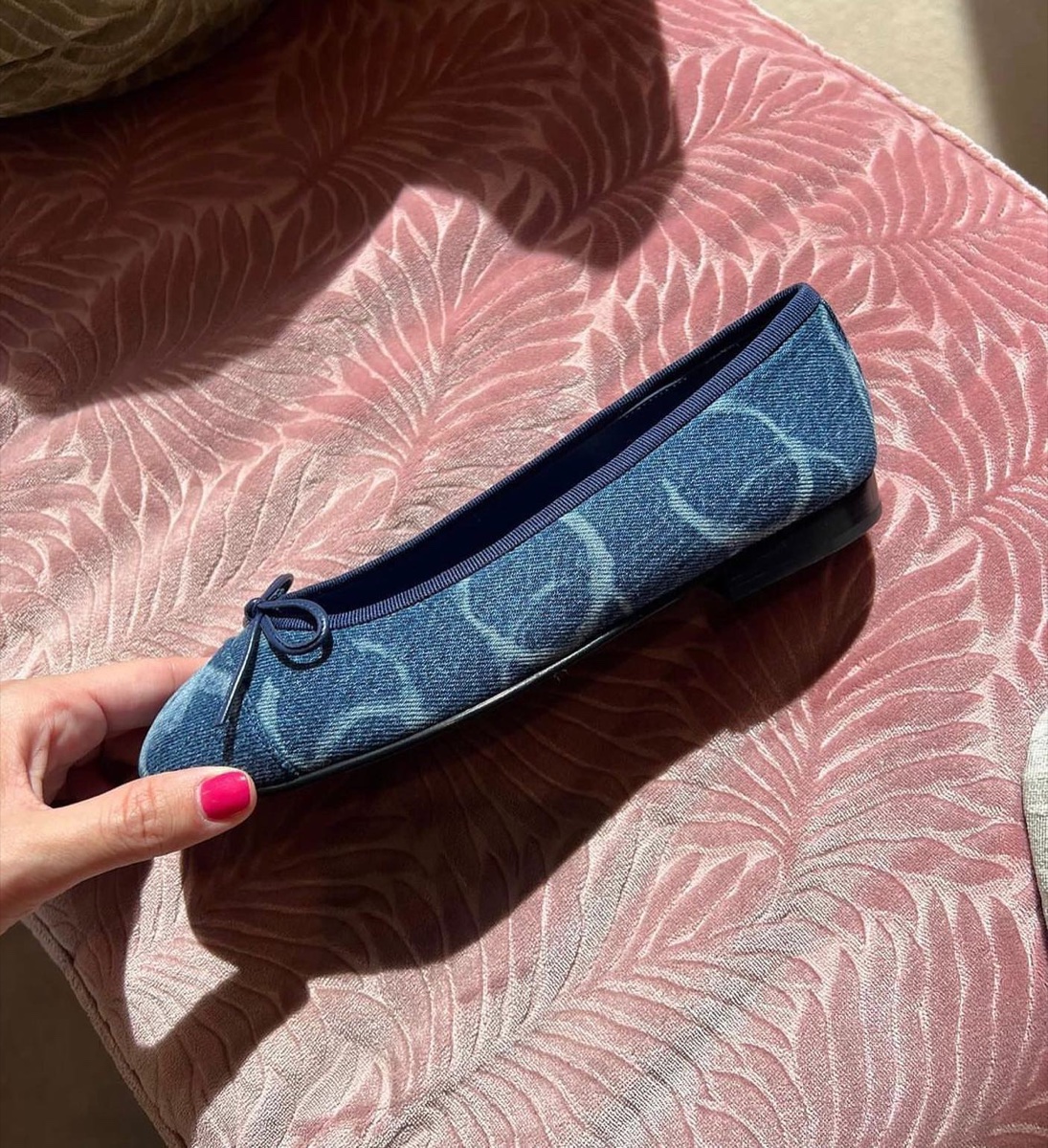 A denim Chanel ballet flat with a navy grosgrain trim and bow, held up in one hand over a pink patterned chair