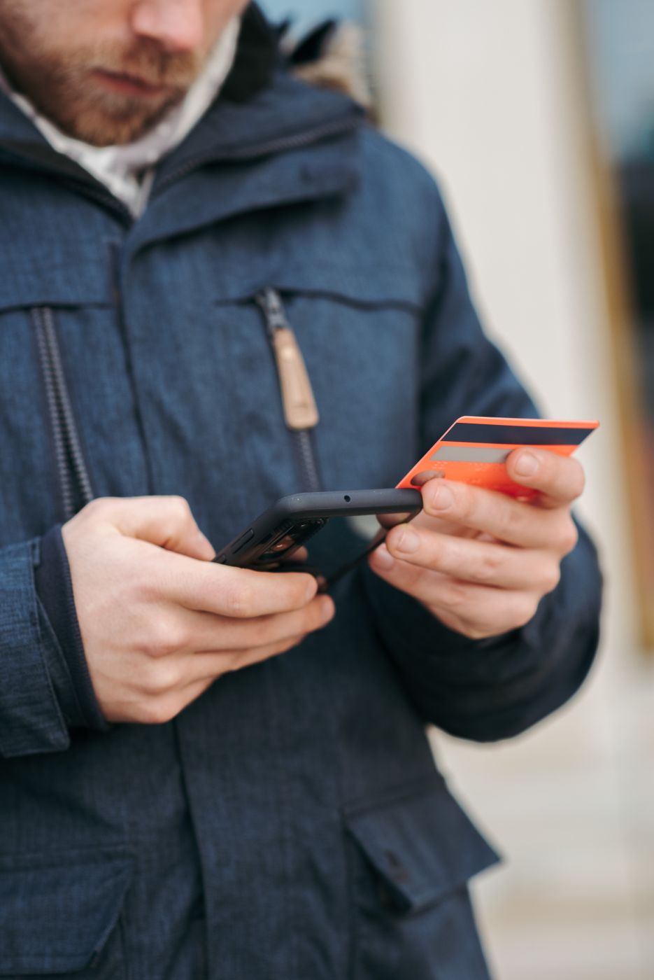 A bearded man in a navy winter parka standing on a street, entering card details into a phone held in his other hand