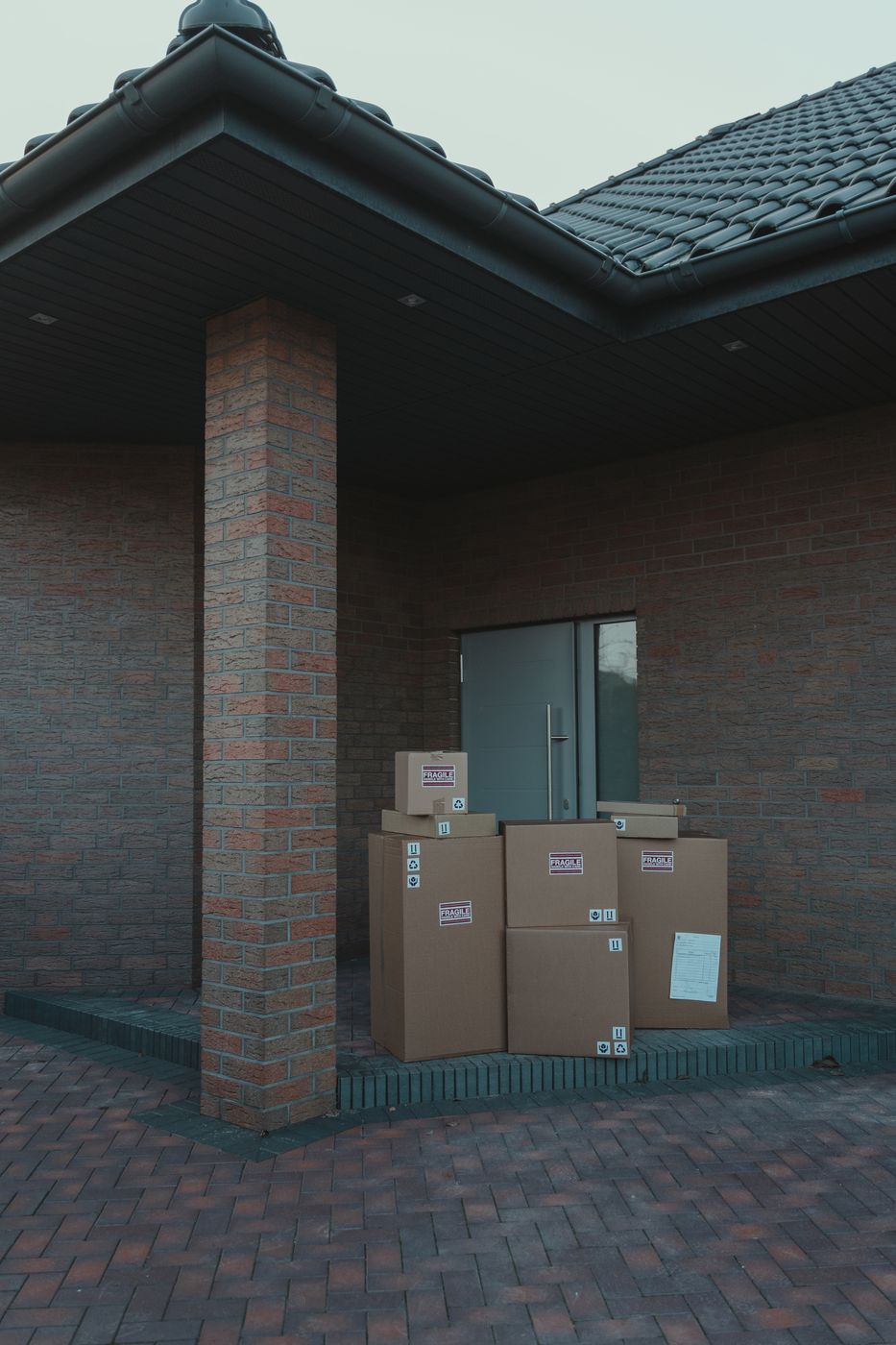 Five plain cardboard parcels of different sizes stacked against a grey door in the brick entryway of a modern house