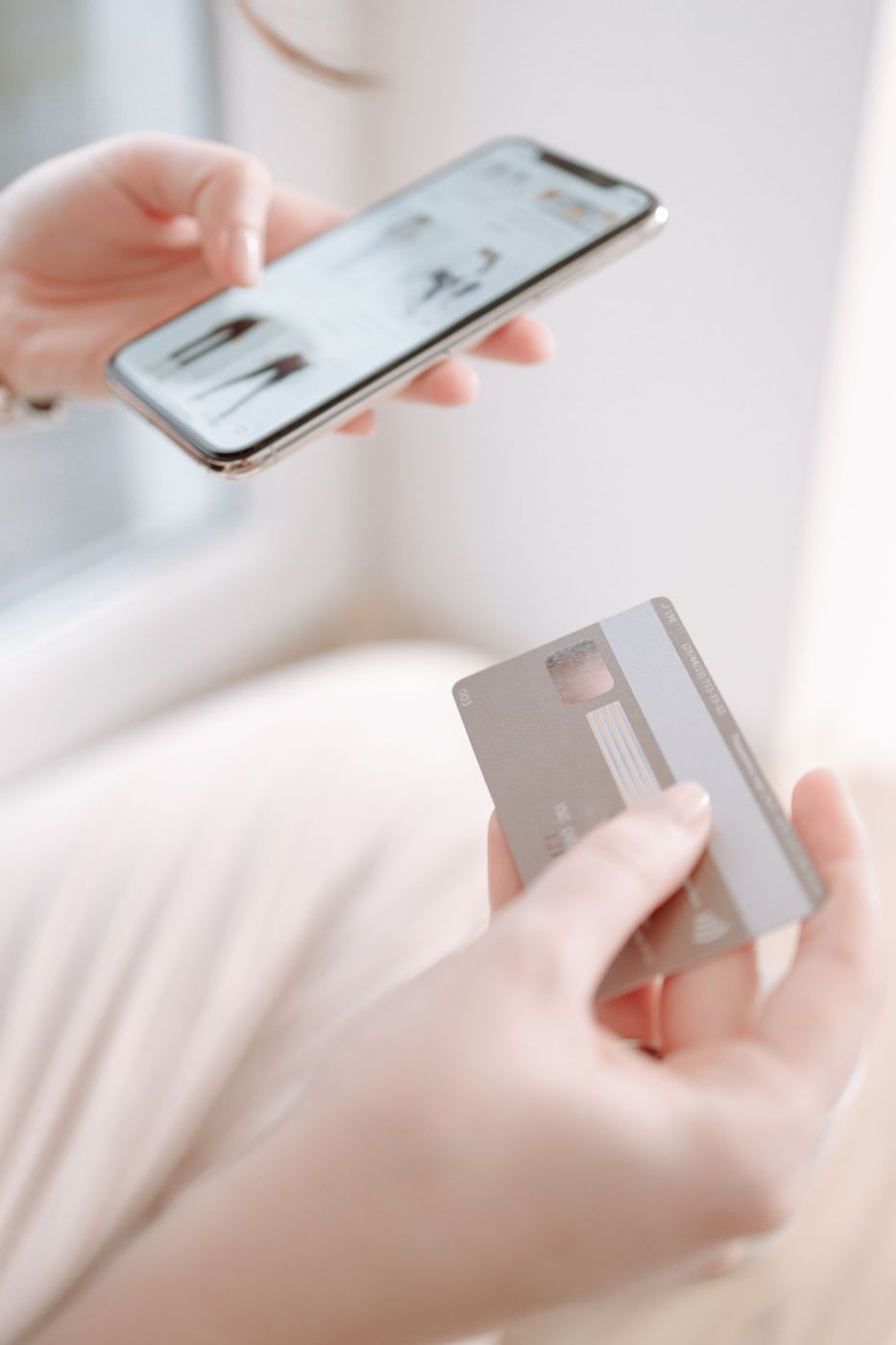 Two hands at a bright window, one holding a smartphone showing a clothing listing page and the other holding a grey payment card