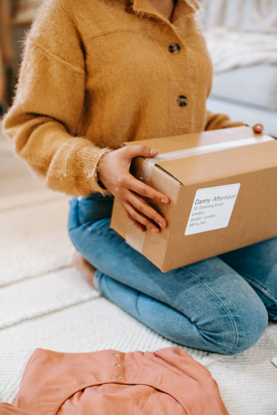 Woman kneeling on the floor with a cardboard parcel on her lap and a coral top laid out nearby