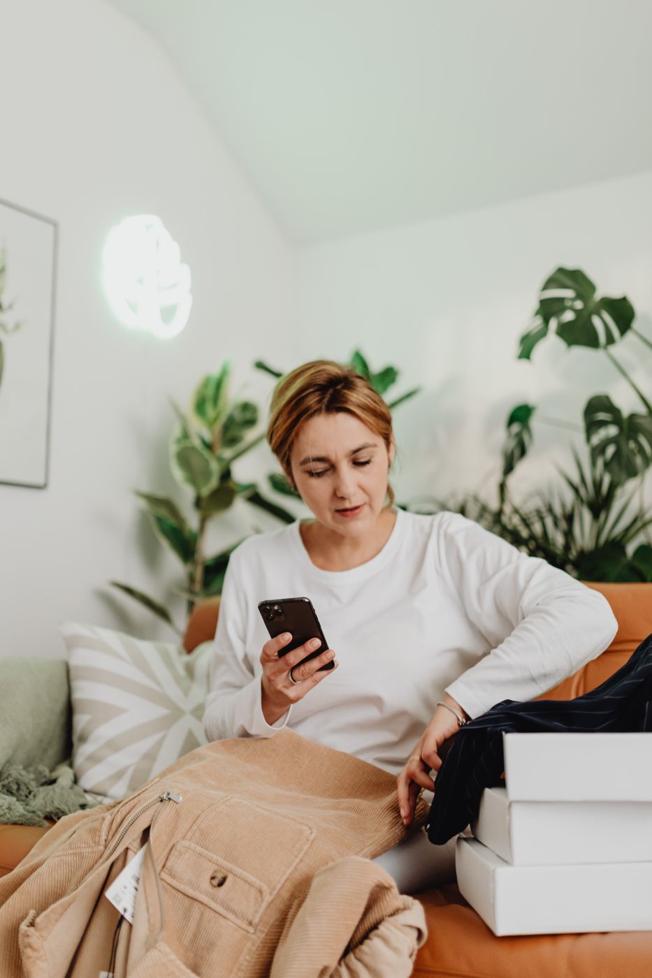 Woman on a sofa checking her phone next to stacked white boxes and a corduroy jacket