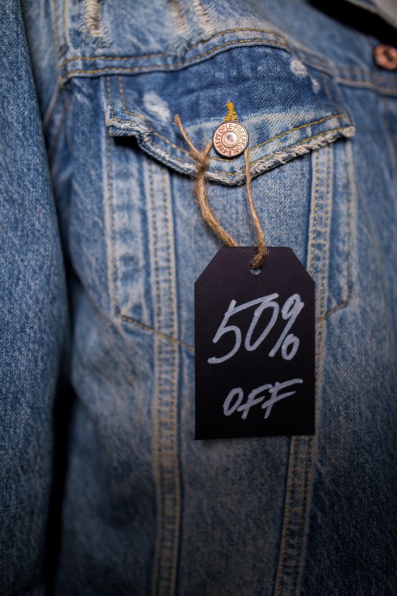 A faded blue denim jacket with a black kraft swing tag tied to the chest pocket, 50% OFF written on it in white marker
