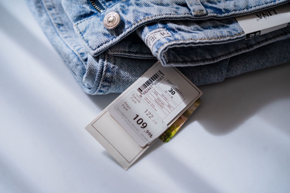 A pair of folded light-wash jeans on a white surface with a printed retail swing ticket showing two prices, 122.21 and 109.99