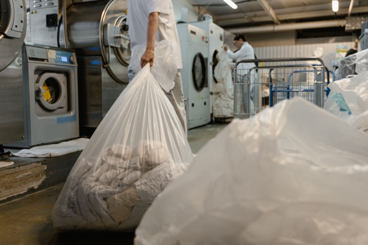 A worker in white carries a large clear plastic bag of bedding across a commercial cleaning floor lined with industrial washers and dryers