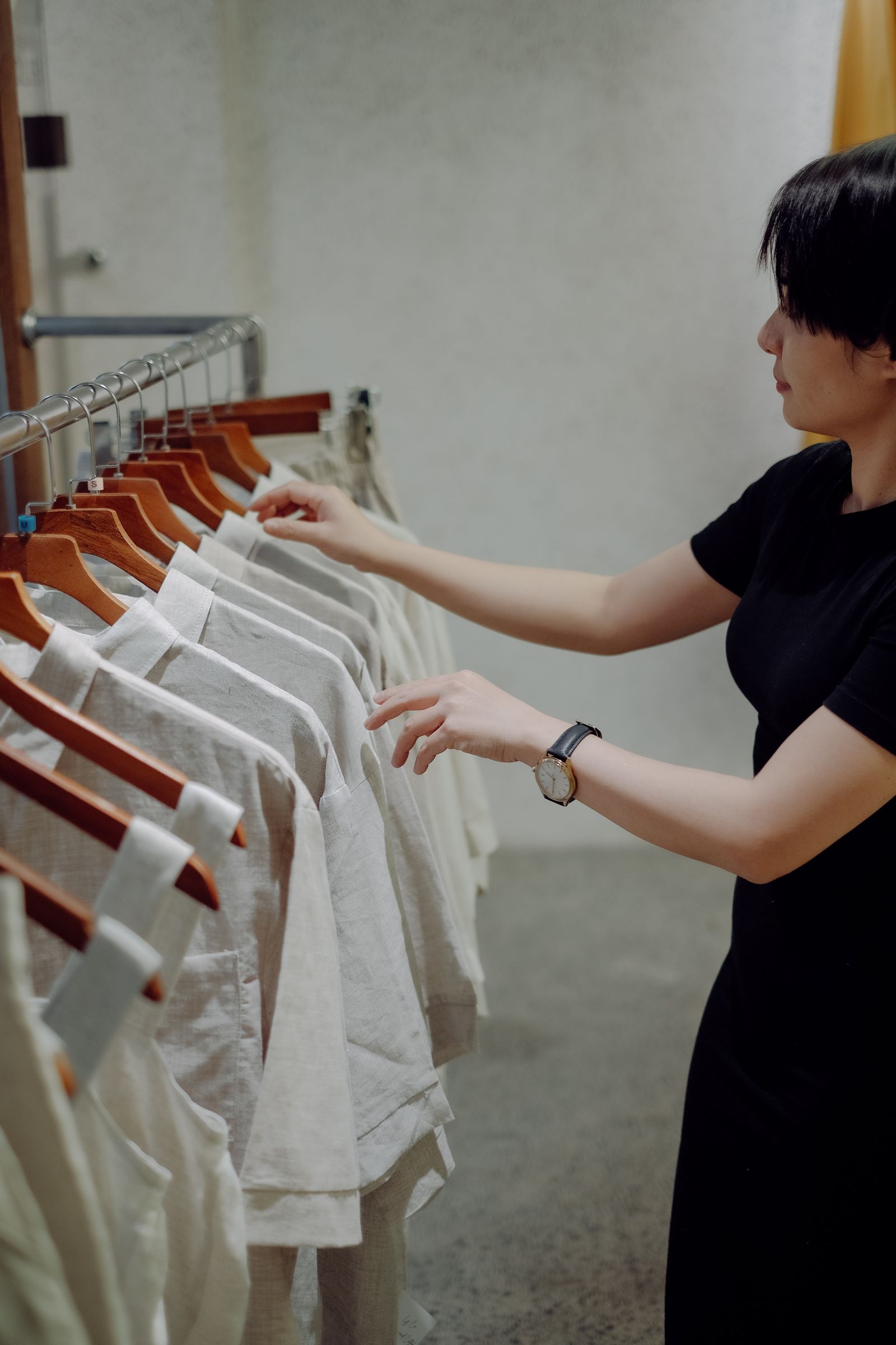 A woman in a black dress reaching into a rail of beige linen shirts on wooden hangers, seen from the side
