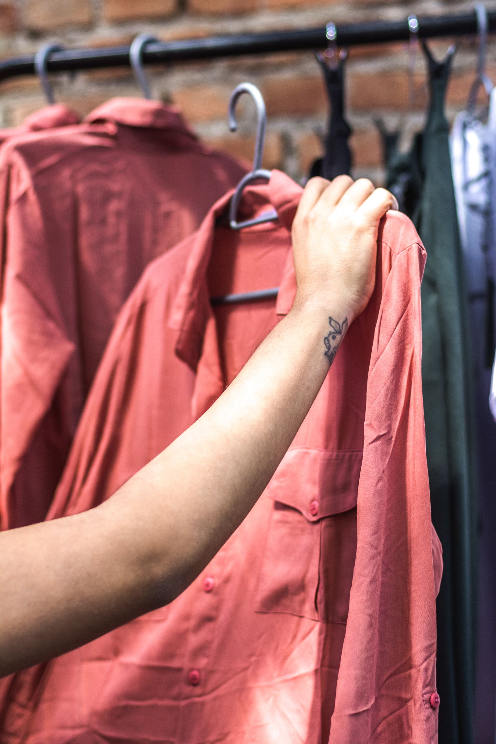 A hand gripping the shoulder of a salmon shirt on a metal hanger, more shirts and a brick wall blurred behind it