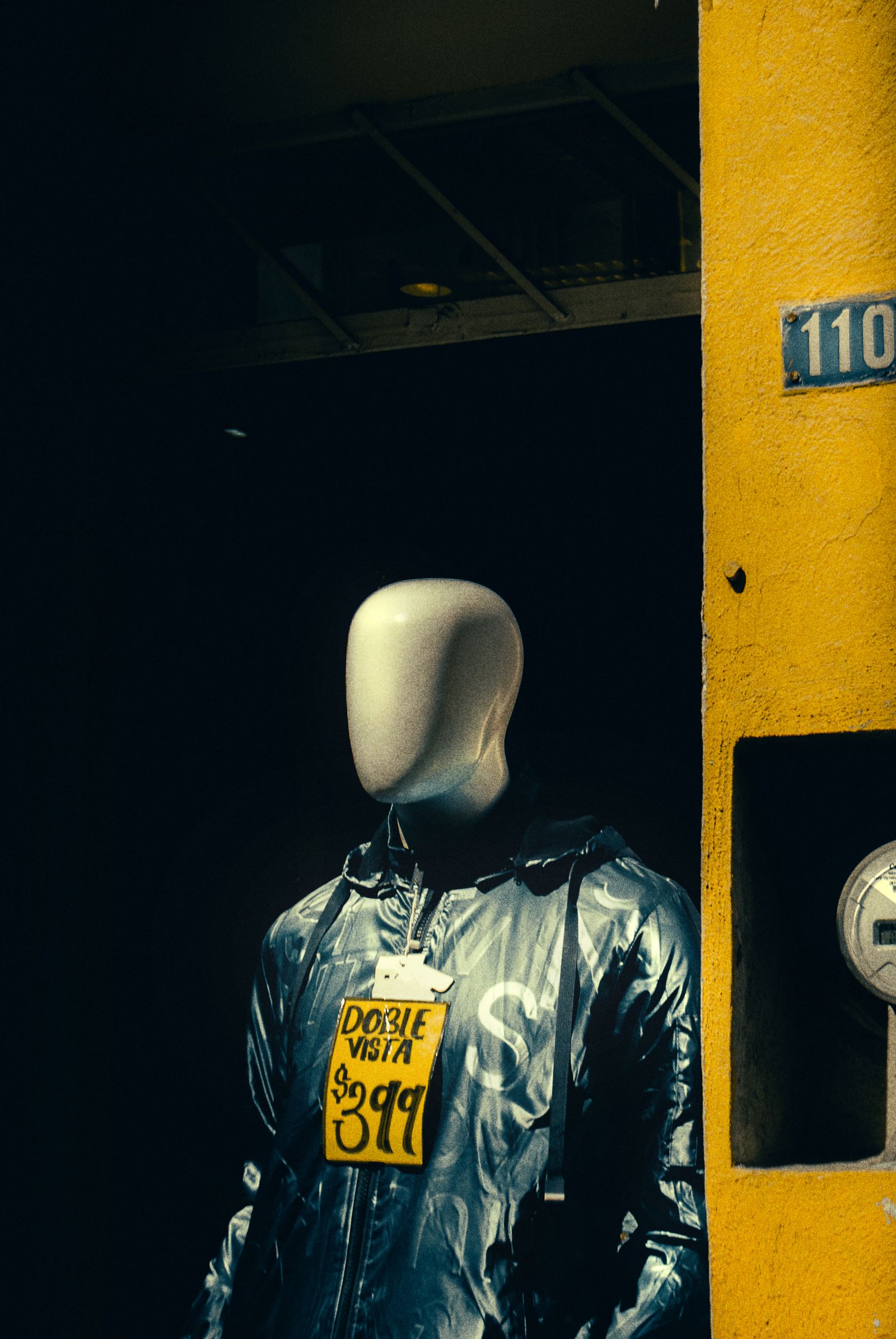 A faceless mannequin in a shiny jacket wearing a handwritten cardboard price sign around its neck in a dark doorway