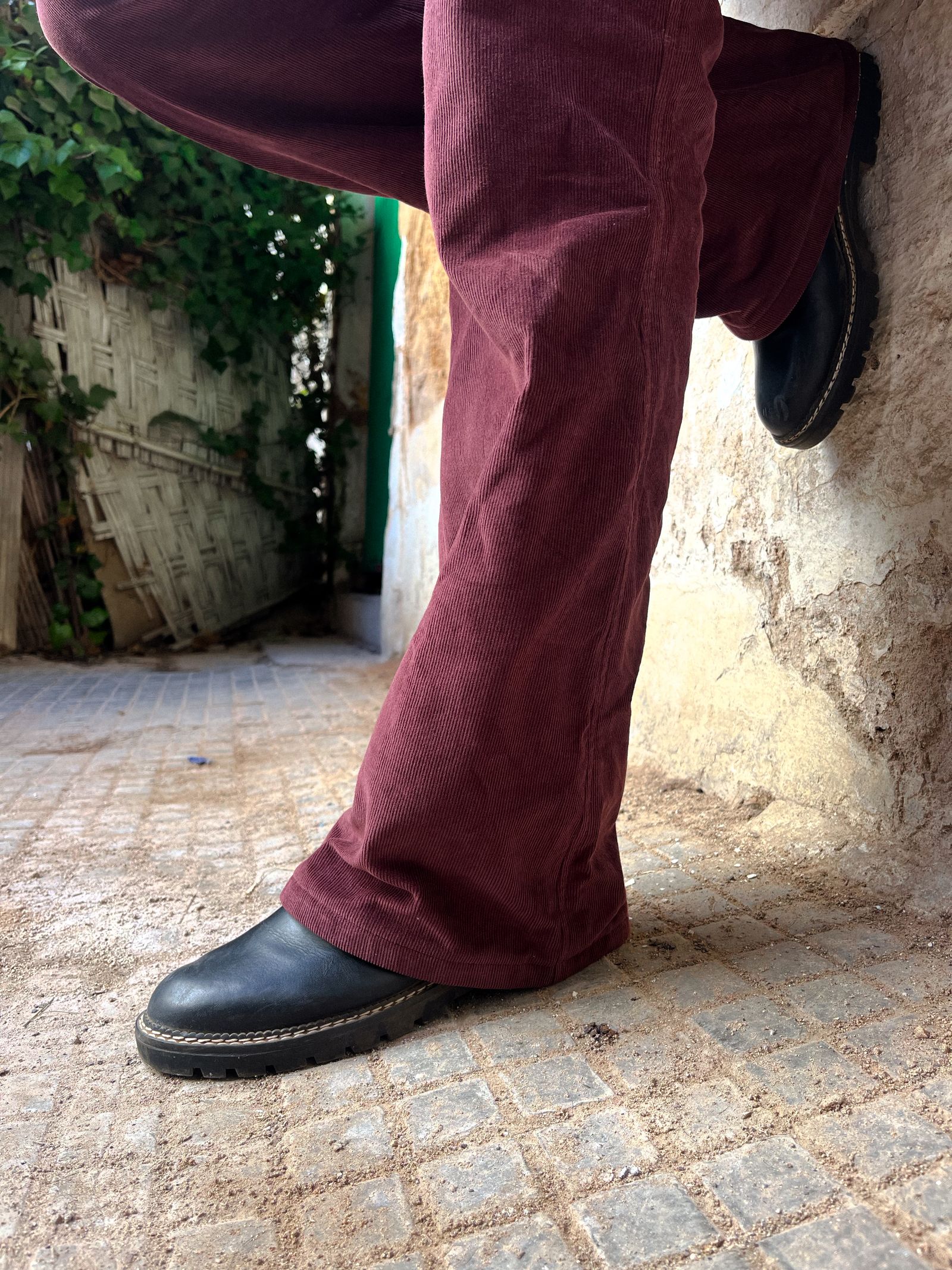 Fine-wale maroon corduroy flares worn with black lug-sole shoes against a stone wall on a cobbled street