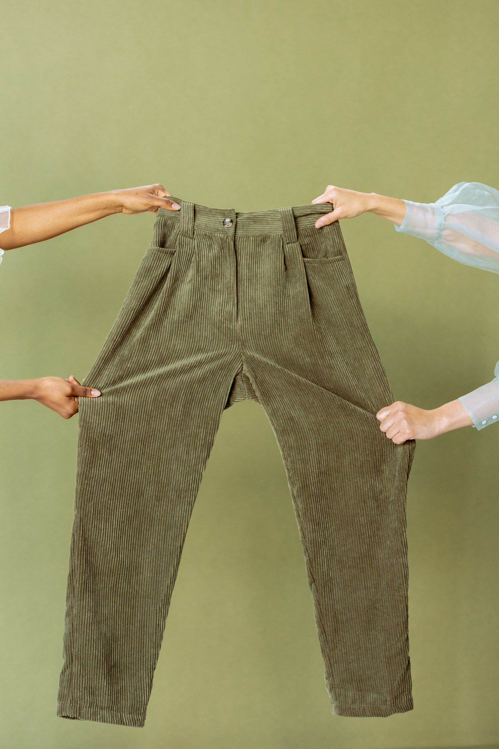 Two pairs of hands holding up a pair of olive green wide-wale corduroy pants against a matching green background