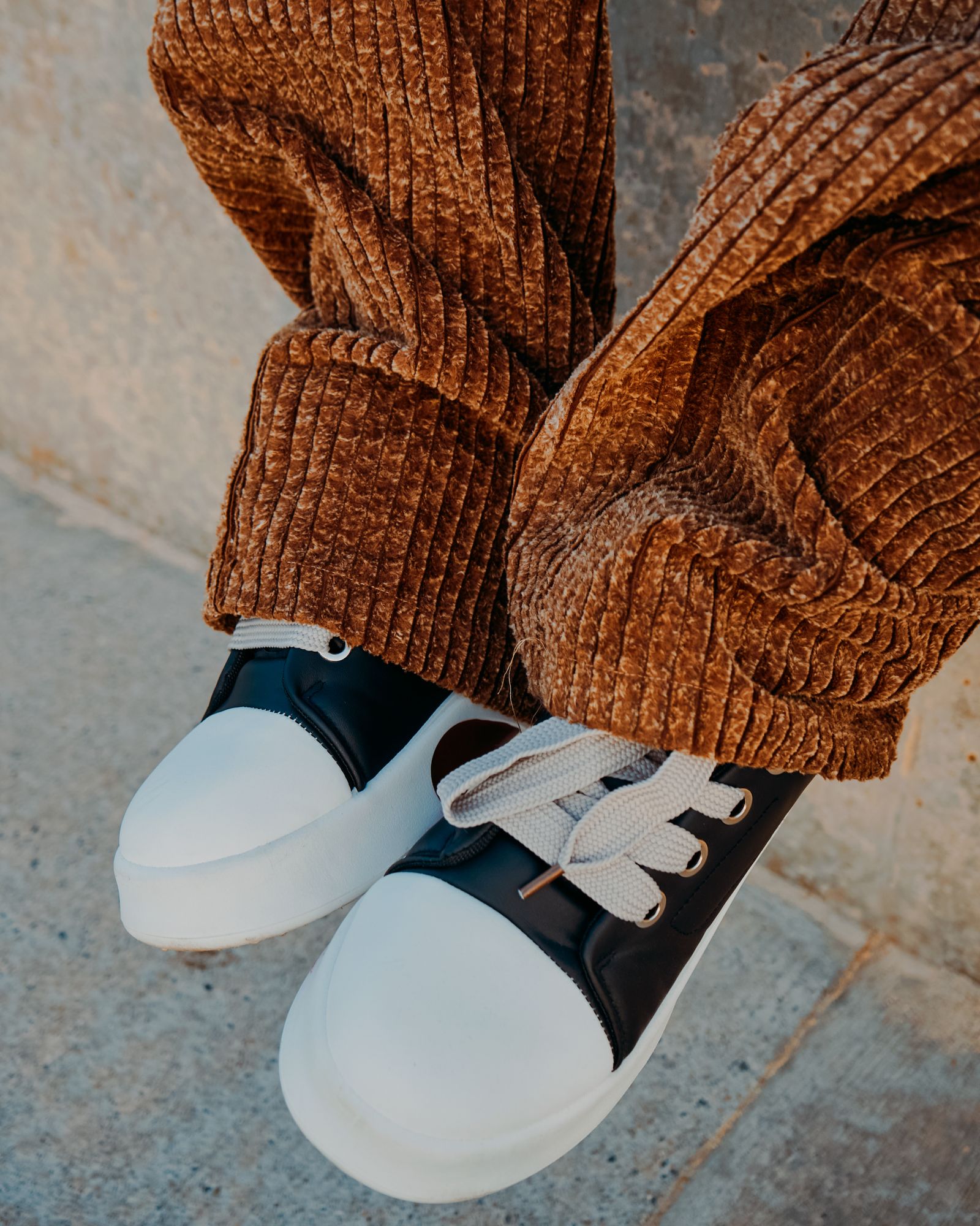 Close-up of chunky wide-wale caramel corduroy pants breaking over black and white sneakers on a concrete step