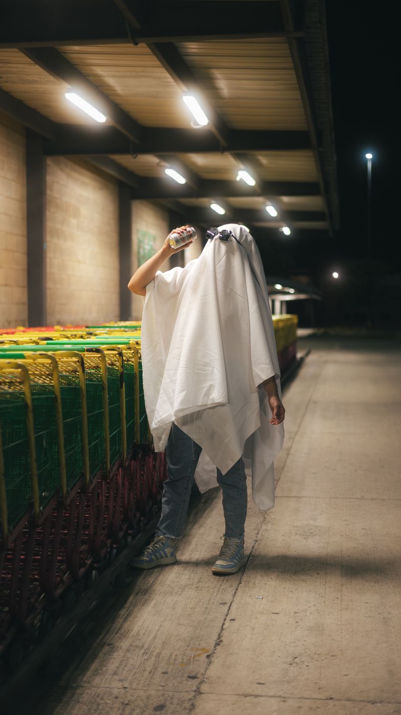 Person in a white sheet ghost costume standing beside a row of shopping trolleys in a store car park at night, drinking from a can