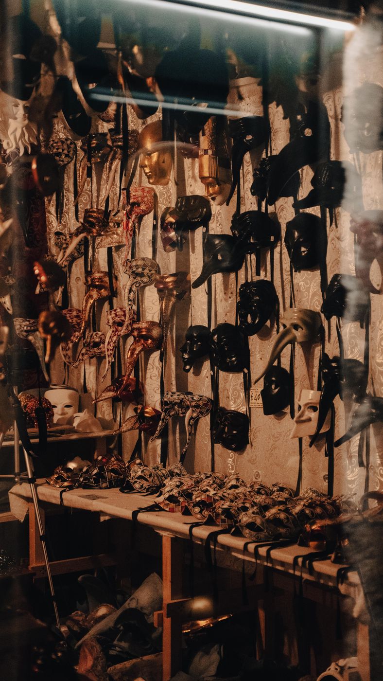 Wall of ornate carnival masks hanging on ribbons above a table of smaller masks in a dim artisan shop