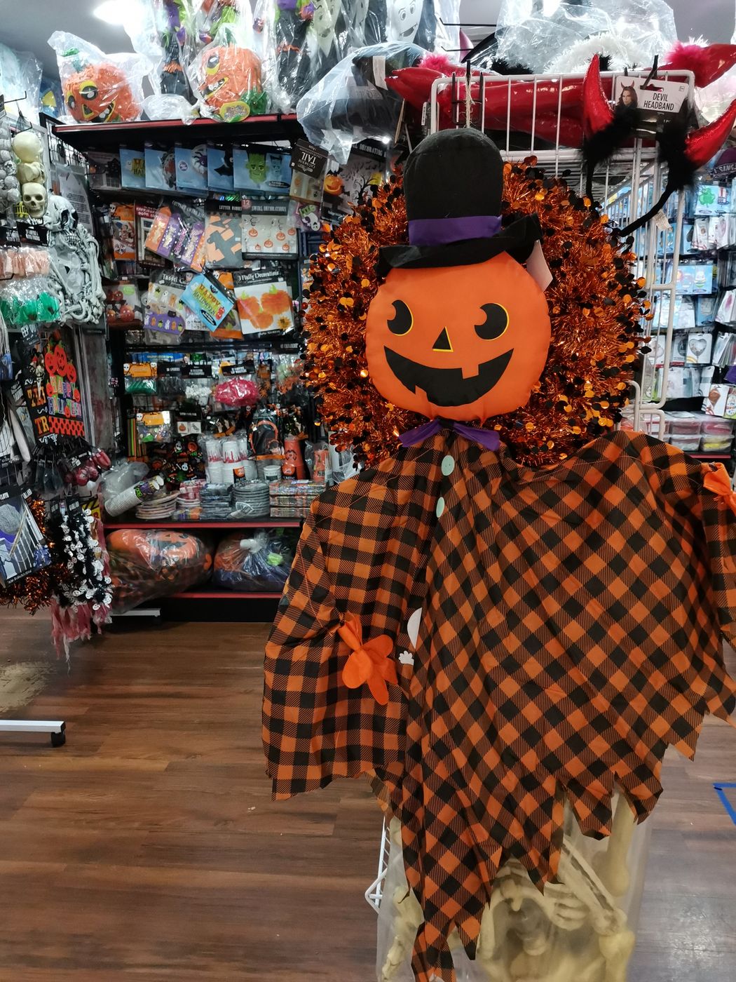 Interior of a costume shop with a pumpkin-headed scarecrow figure in orange plaid standing in front of pegboard walls hung with packaged masks, wigs and Halloween accessories