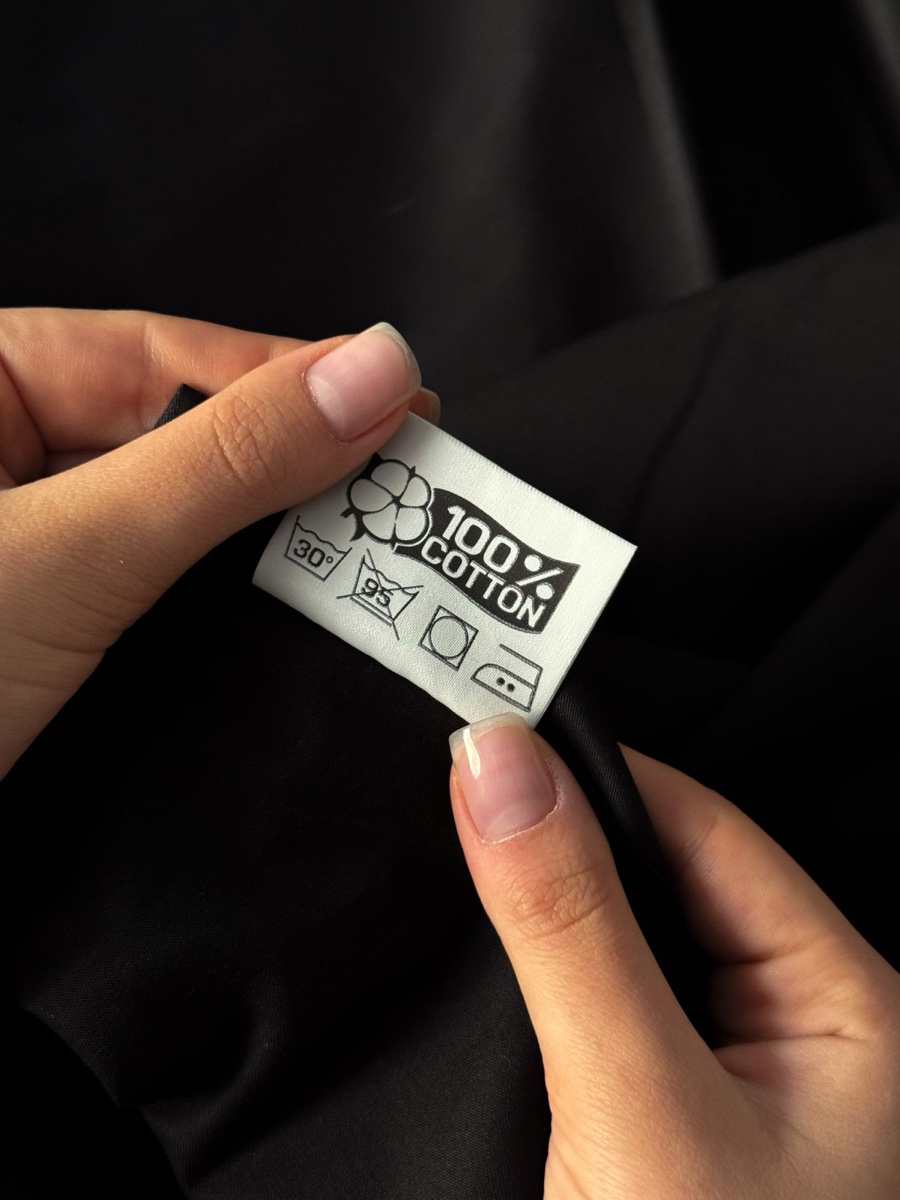 Two hands holding a white sewn-in garment label printed with a cotton flower symbol, 100% cotton, and wash care icons against black fabric