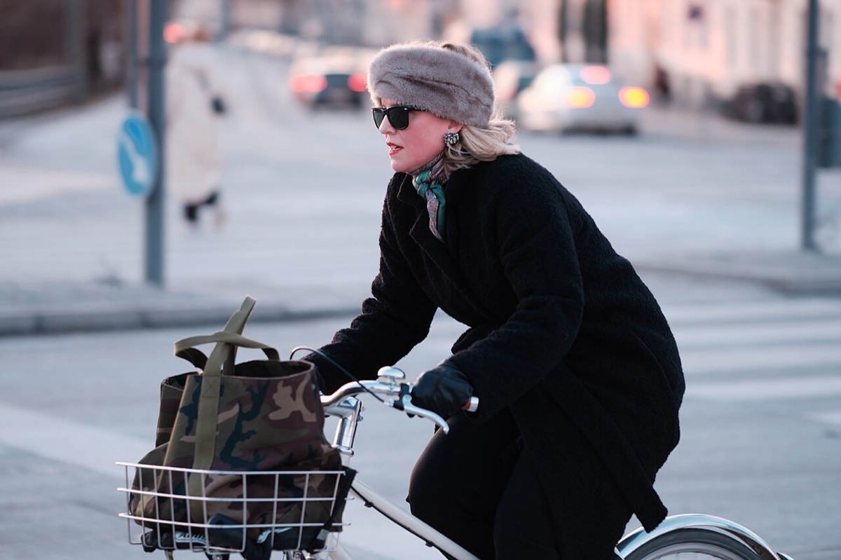 A woman in a black bouclé coat, printed scarf and gray fur headband cycles through a Copenhagen intersection with a camouflage tote in her basket