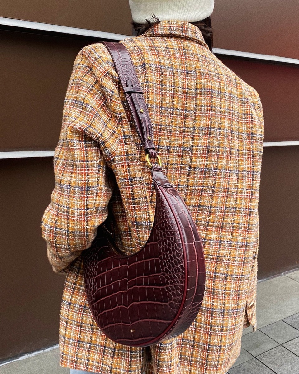 Seen from behind, a person in a rust plaid wool blazer carries a burgundy croc-embossed half-moon shoulder bag on a brown street
