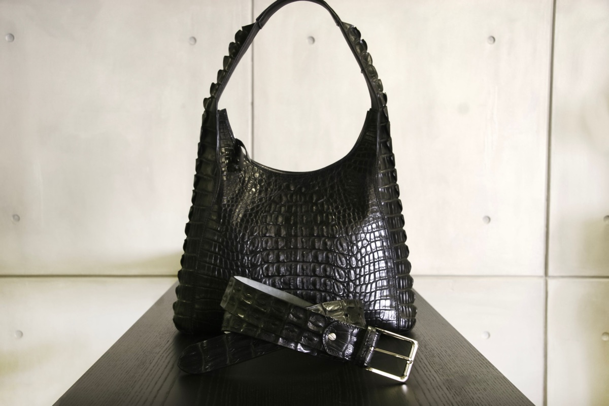 A black handbag and matching belt covered in crocodile scale texture, displayed on a dark table against a pale tiled wall