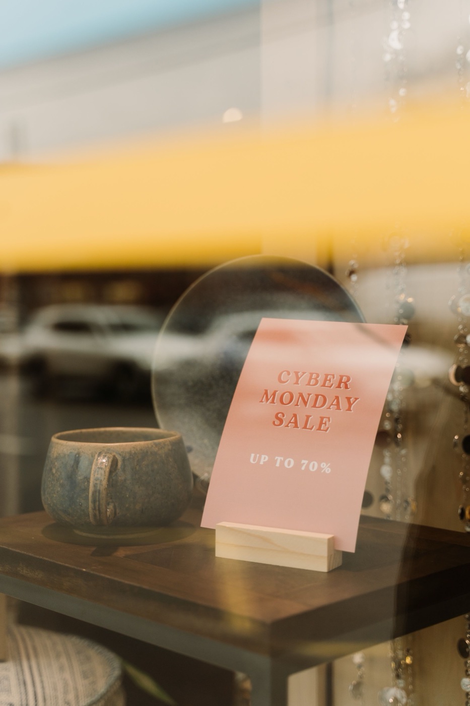 A pink Cyber Monday sale sign reading up to 70 percent off propped on a small wooden stand in a shop window beside a ceramic mug