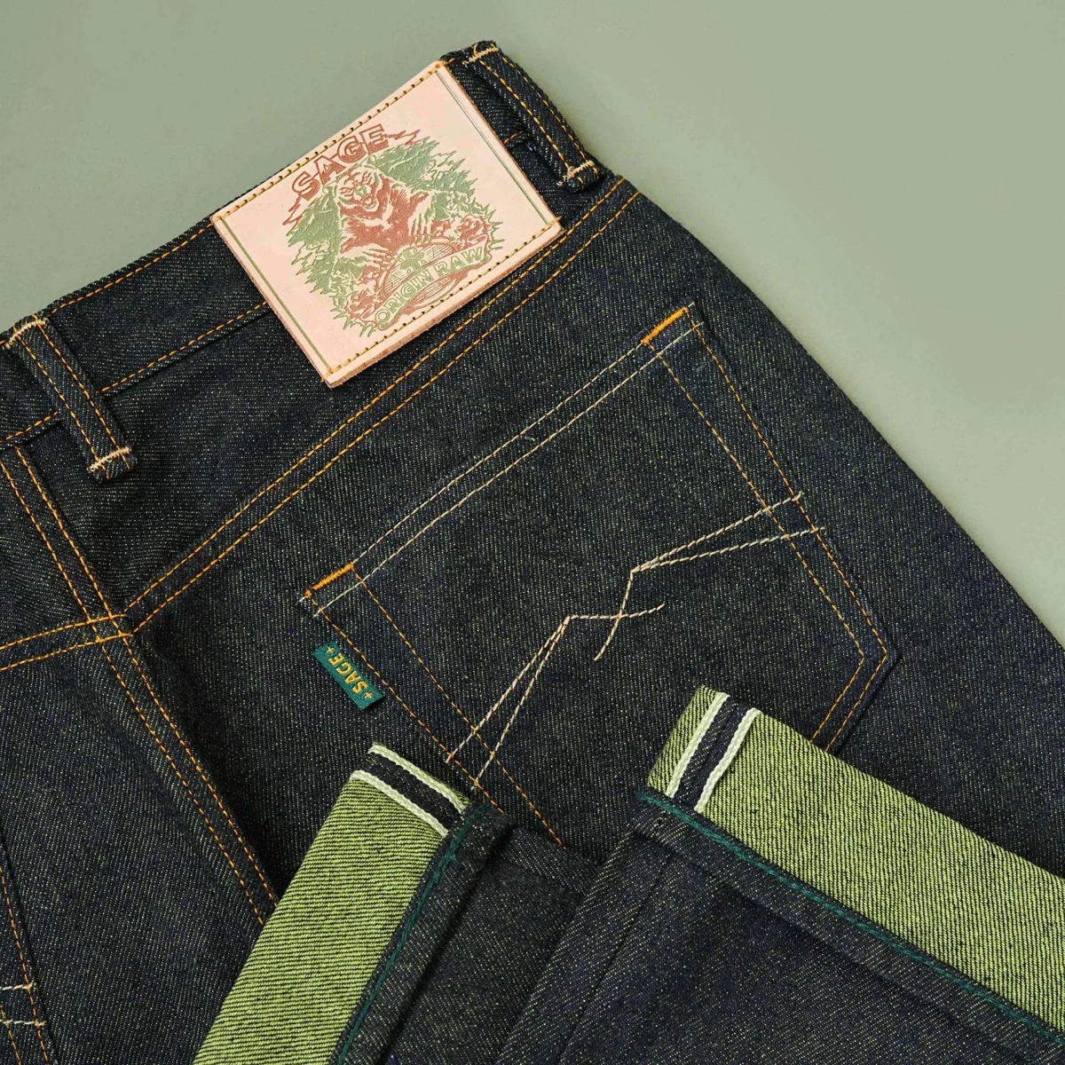 Close-up of the back pocket and cuffed selvedge hem of a pair of raw deep indigo jeans with a leather patch, on a sage background
