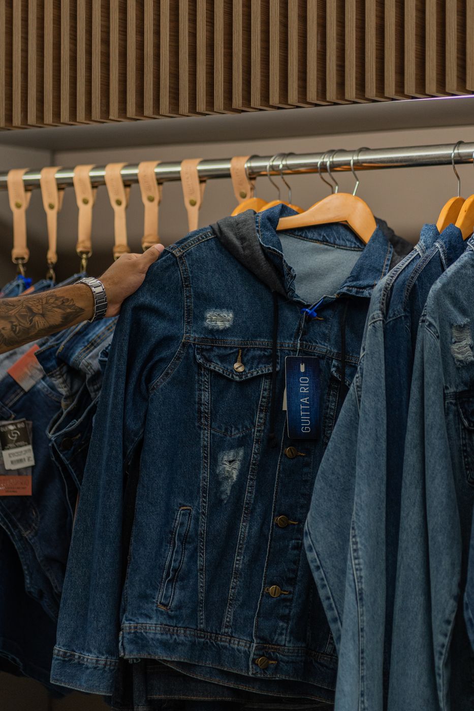 A hand lifting one denim jacket forward on a rail of near-identical denim jackets, brand hang tags visible on the fronts