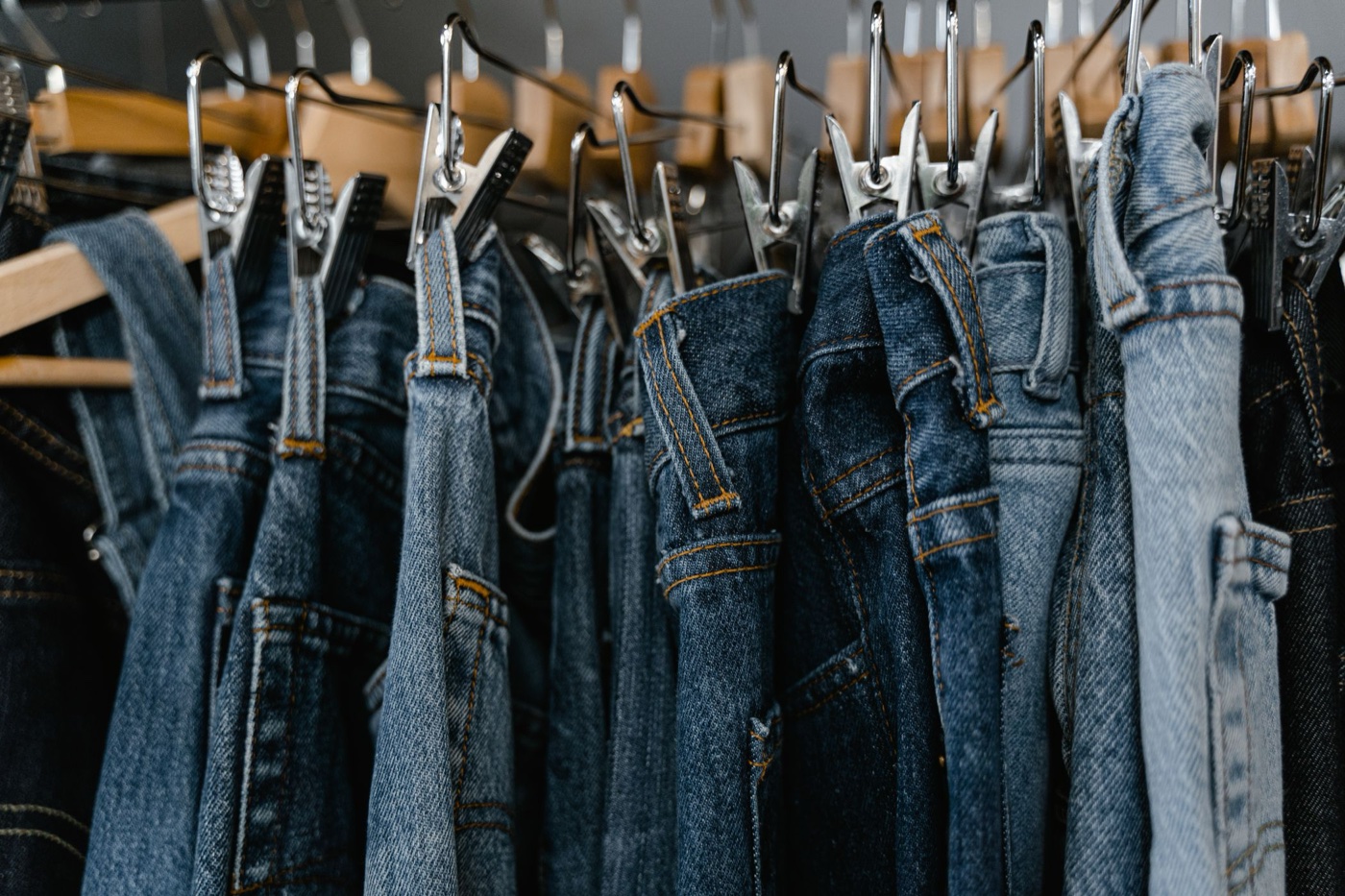 A close row of blue jeans clipped by their waistbands to metal hangers on a store rail, washes ranging from pale to dark indigo
