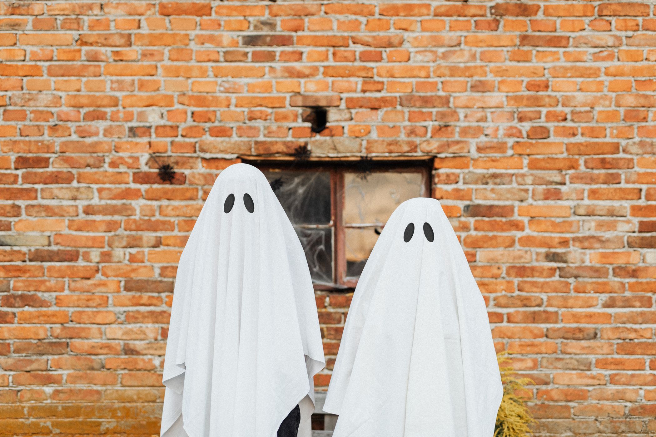 Two people wearing white bedsheet ghost costumes with cut-out eyes standing in front of a brick wall