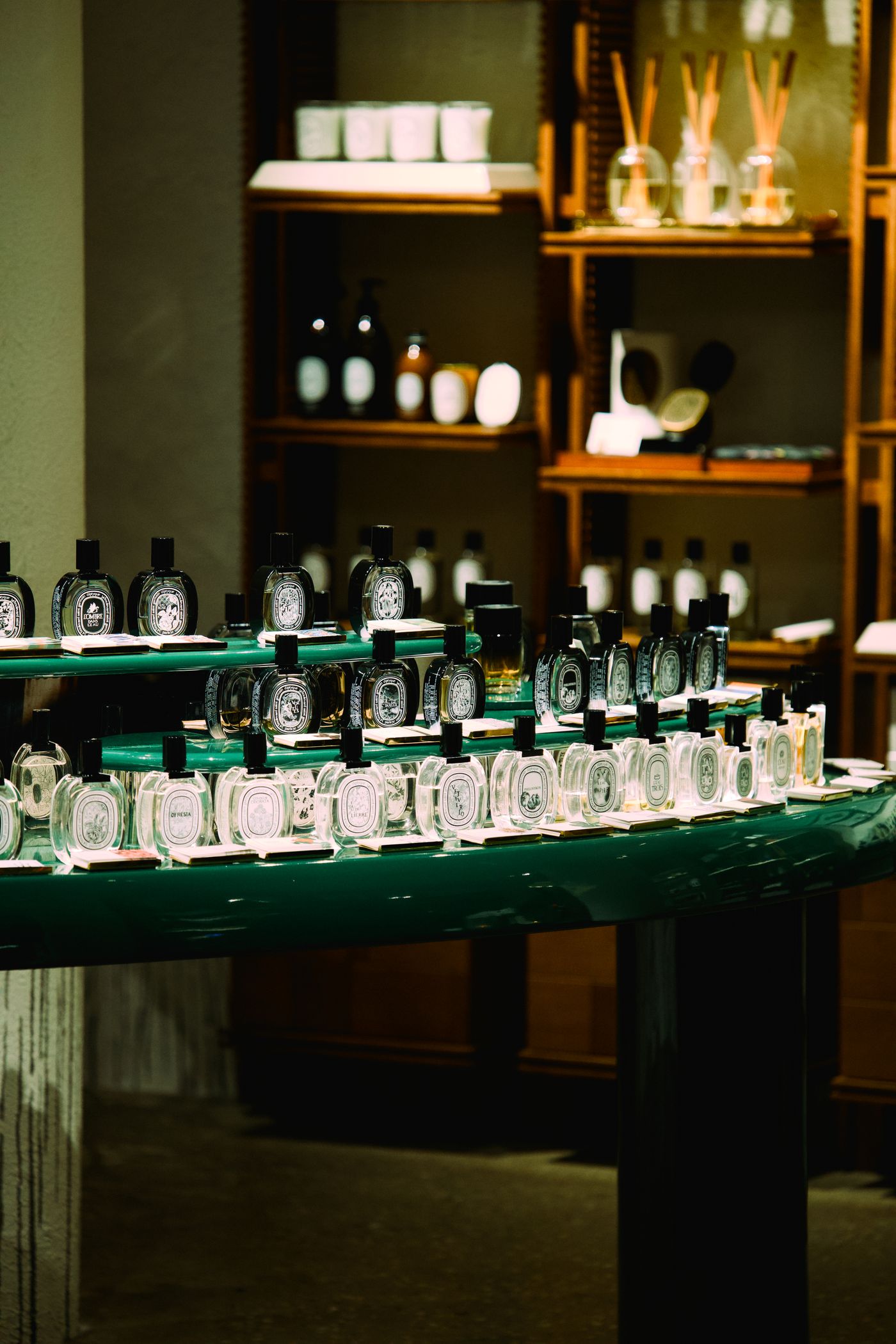 A green tester table holding rows of oval perfume bottles in a dim boutique, with candles and diffusers shelved behind