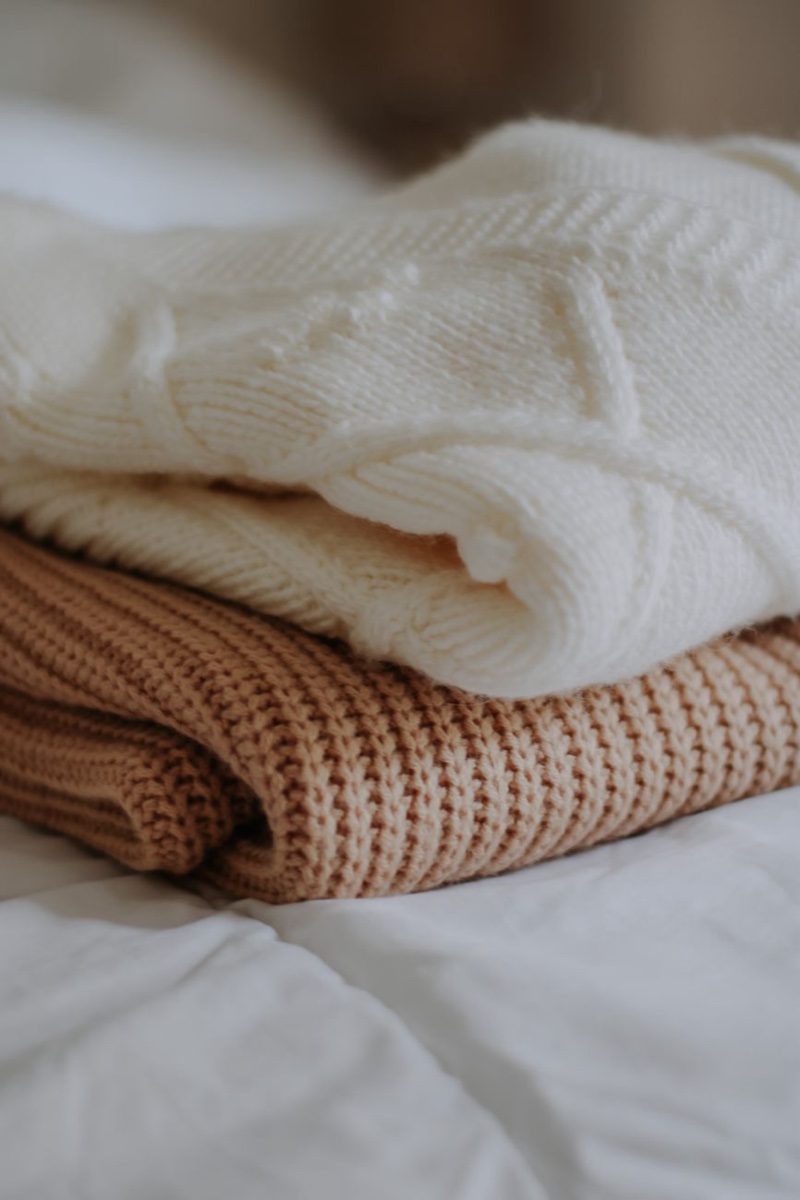 Two folded knit sweaters, one cream and one camel, stacked on a white bed
