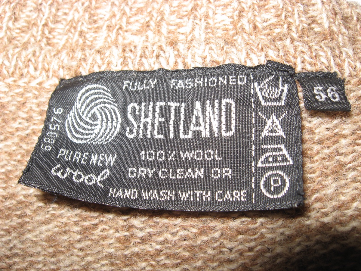 A black woven care label sewn into a camel Shetland sweater reading Pure New Wool, Fully Fashioned, 100% Wool, Dry Clean or Hand Wash With Care, with care symbols along the right edge