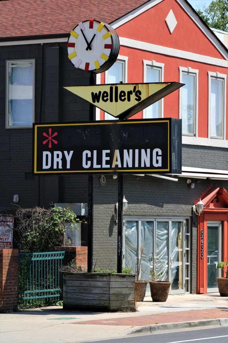 A yellow and black Weller's Dry Cleaning sign with a clock mounted above it on a brick storefront