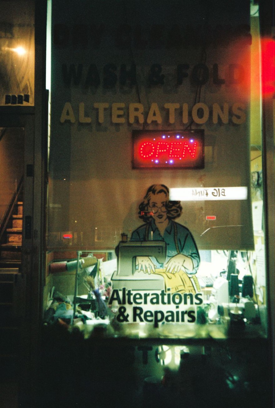 Night view through a shop window lettered DRY CLEANING, WASH AND FOLD, ALTERATIONS with a red neon OPEN sign and a sewing machine decal