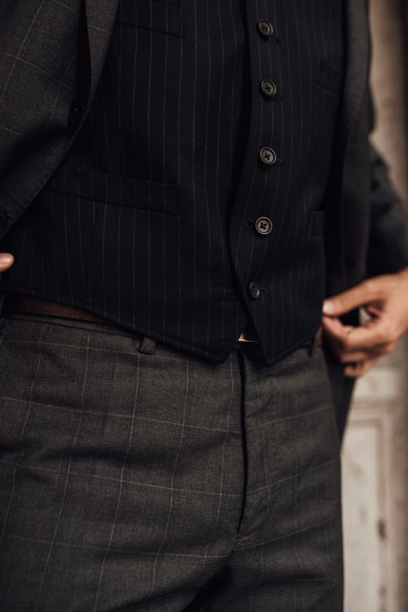 Close-up of grey checked wool suit trousers and a pinstriped waistcoat, hands adjusting the hem of the jacket, no face in frame