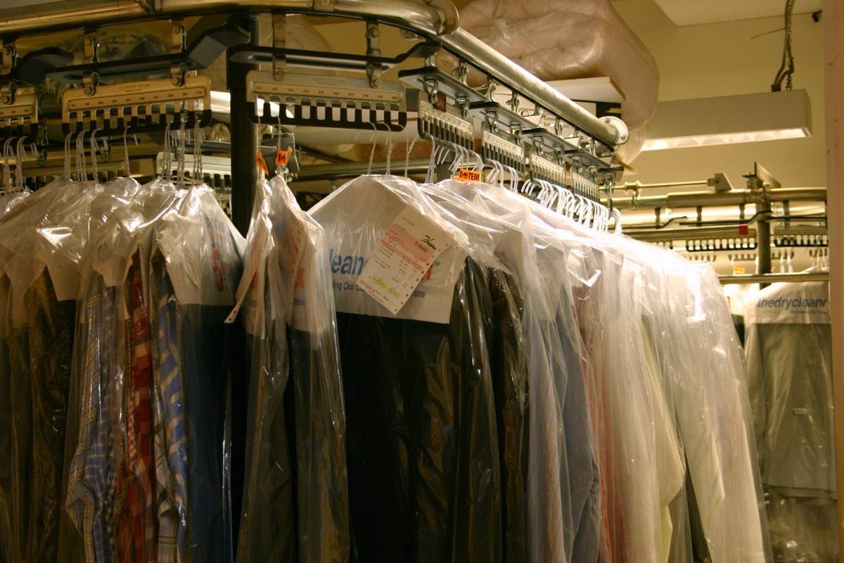 A dry cleaner's overhead conveyor rail loaded with garments in clear plastic bags, a paper ticket stapled to one of them