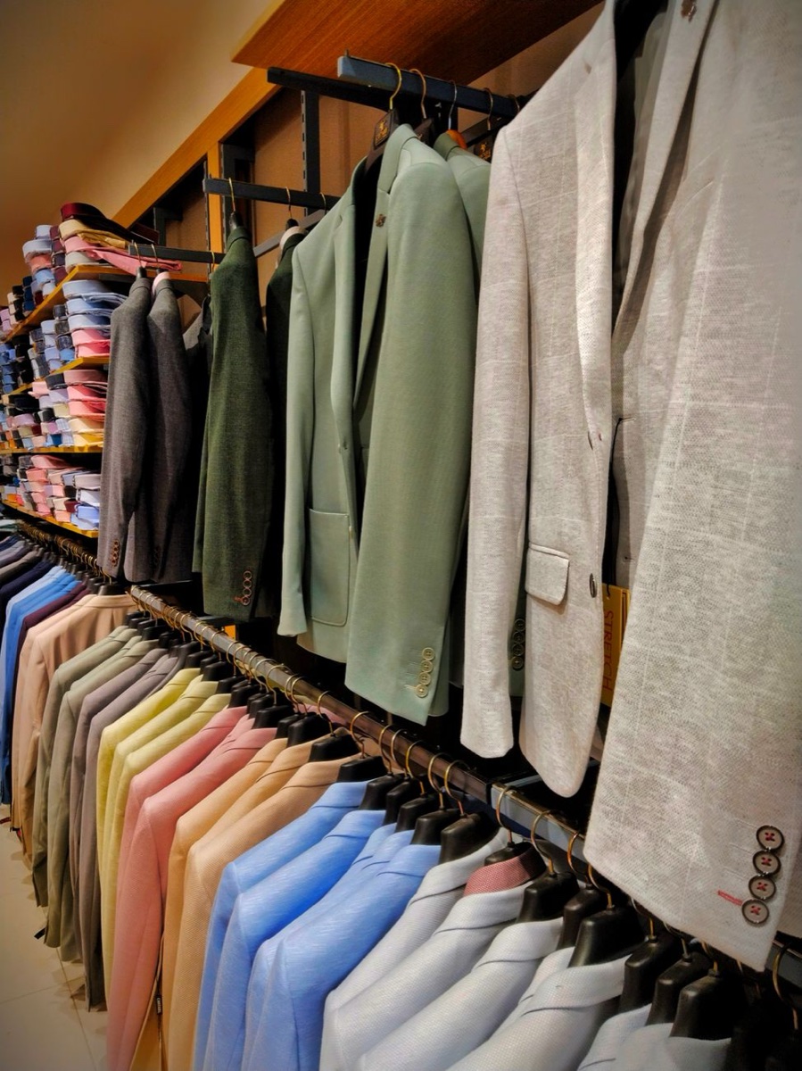 A wall of suit jackets and blazers in greens, greys and pale linen hanging in rows above a rail of folded shirts in a menswear shop