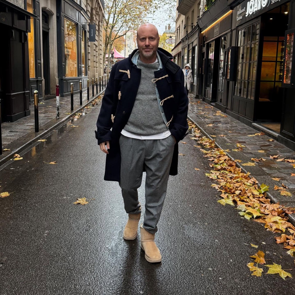 A man in a navy duffle coat with rope toggles over a grey sweater and trousers, walking a wet Paris street scattered with autumn leaves