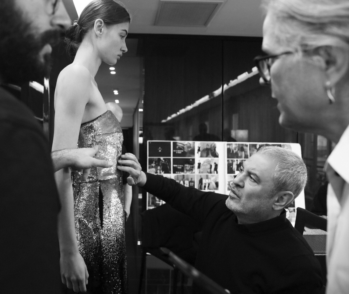 A black-and-white photo of Elie Saab seated, reaching up to adjust a sequinned strapless dress on a model while two colleagues look on