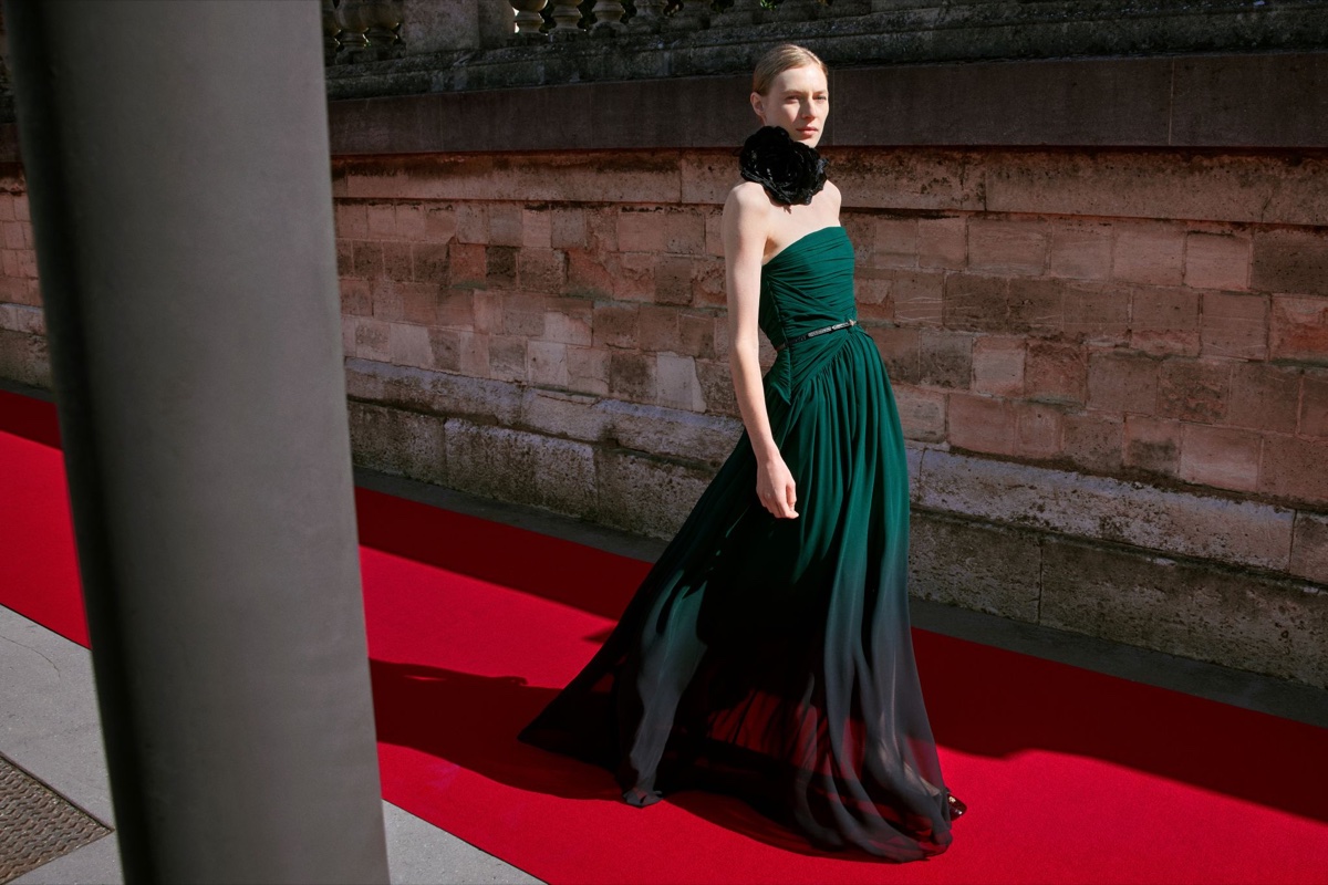 A model stands on a red carpet beside a stone wall wearing a strapless deep-green gown that fades to black at the hem, with a large black flower at her neck