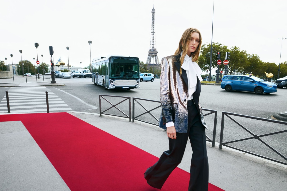 A model in a sequinned blazer, white pussy-bow blouse and black trousers walks past a red carpet laid across a Paris intersection, with the Eiffel Tower and a city bus behind