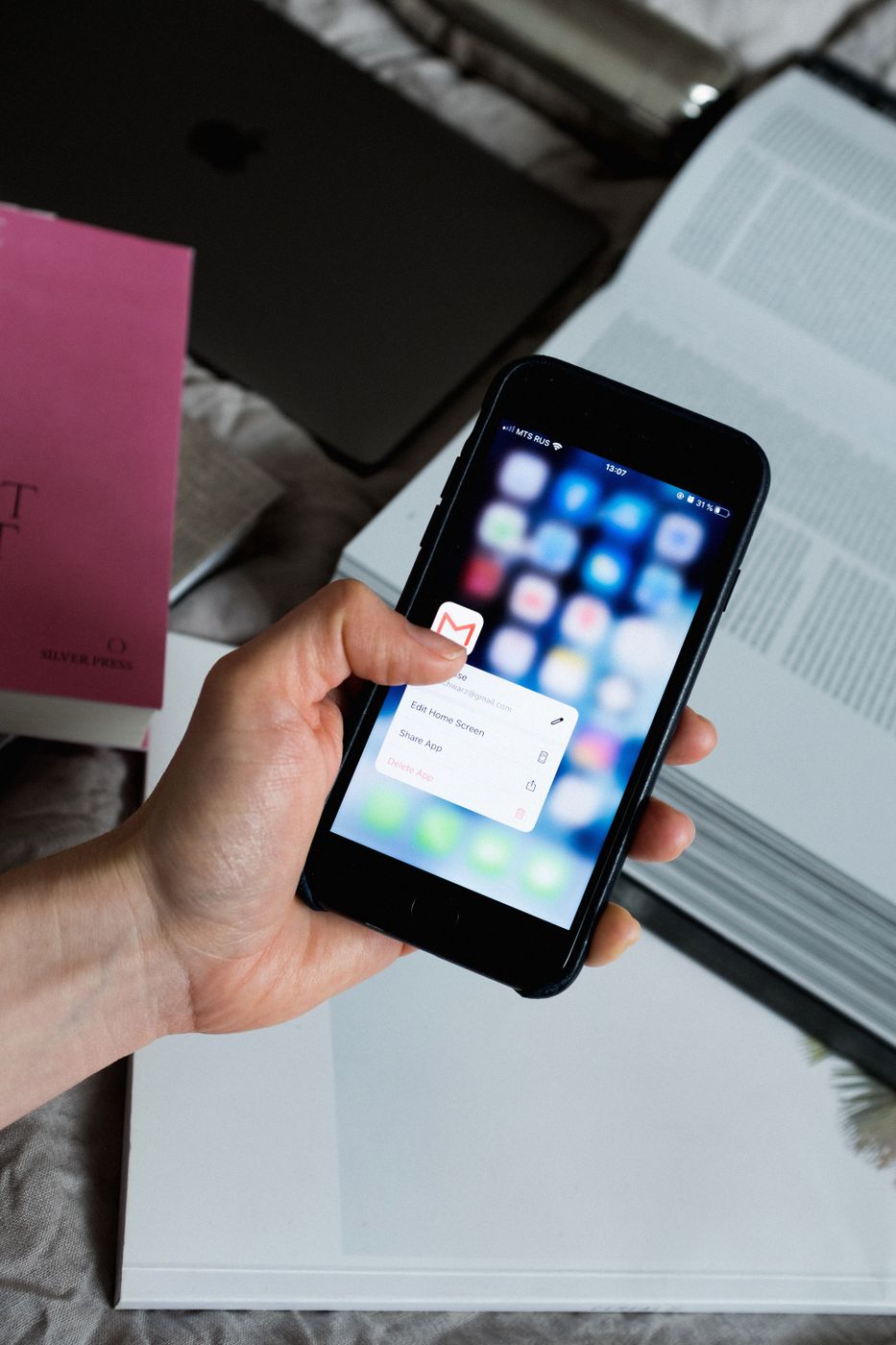 A hand holding a smartphone with a mail app icon pressed on the home screen, books and a laptop in the background