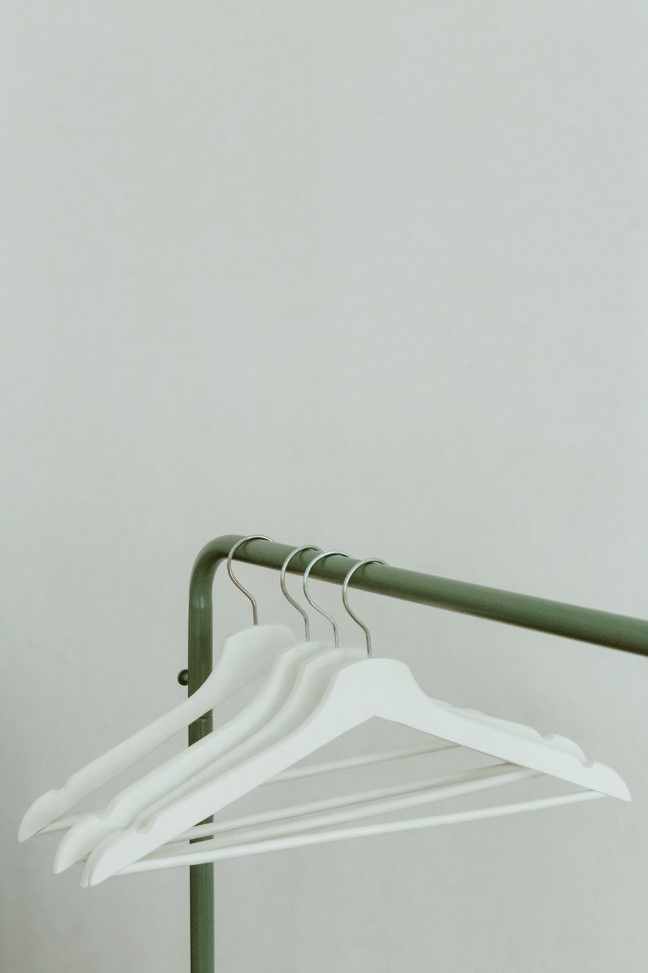 Four bare white hangers left on a green metal clothing rail against a plain off-white wall
