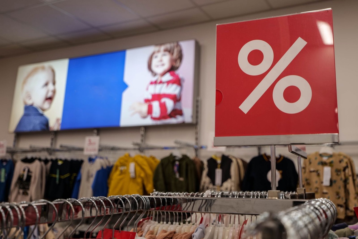 A red square placard printed with a large white percent symbol stands above a rail of children's clothes in a store