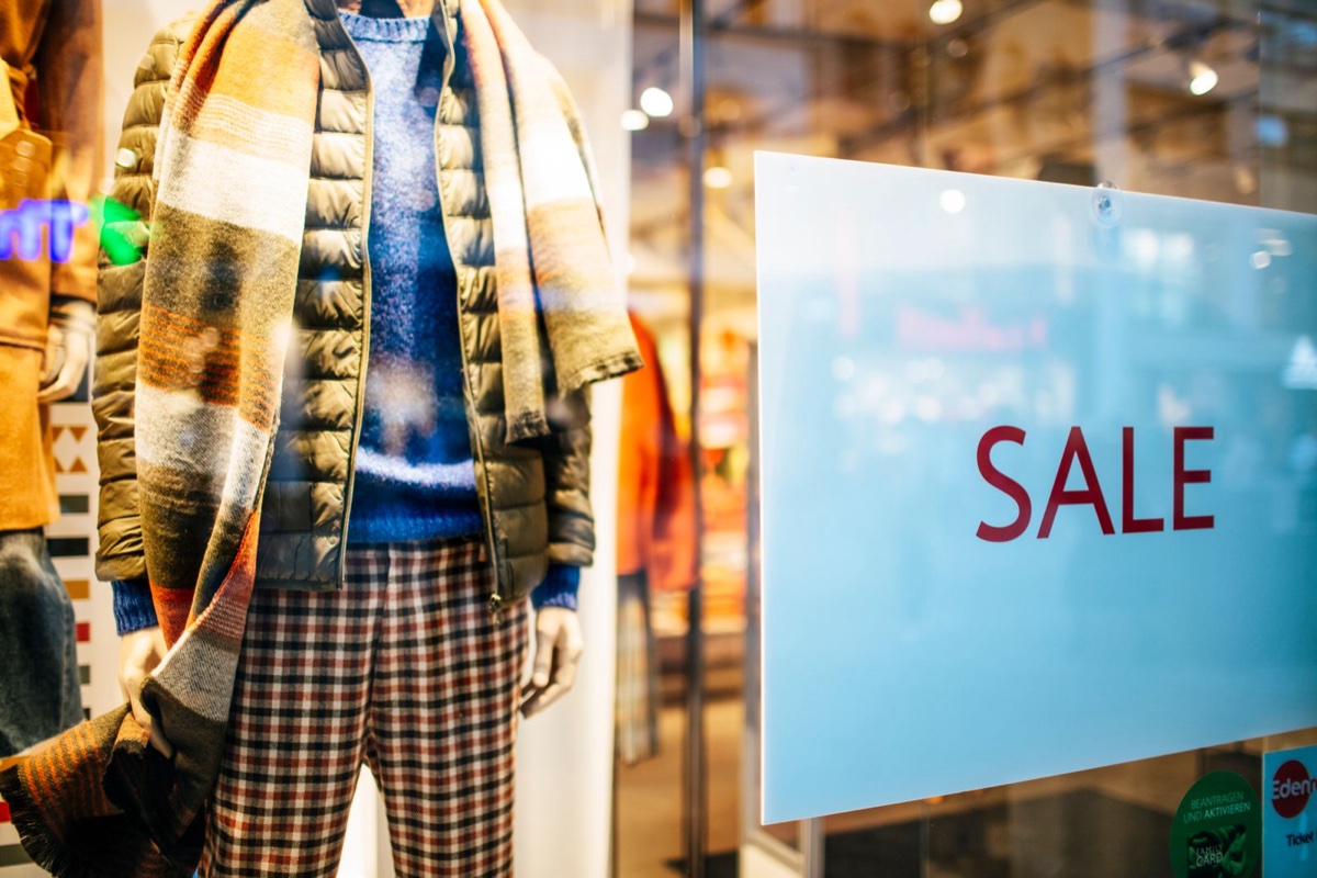 A pale blue SALE card suction-cupped to a store window beside a mannequin dressed in a puffer vest, checked scarf and plaid trousers
