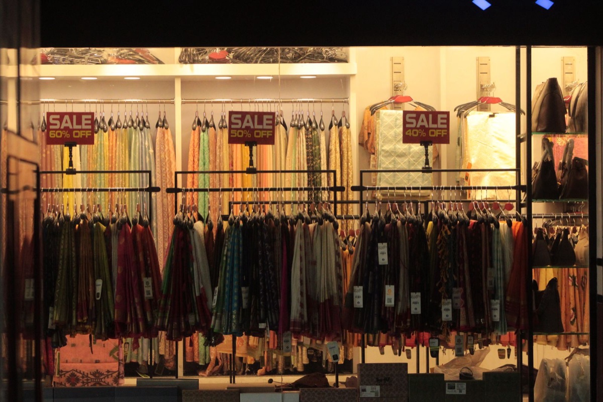 A clothing store window at night with three red placards over separate racks reading SALE 60% OFF, SALE 50% OFF and SALE 40% OFF