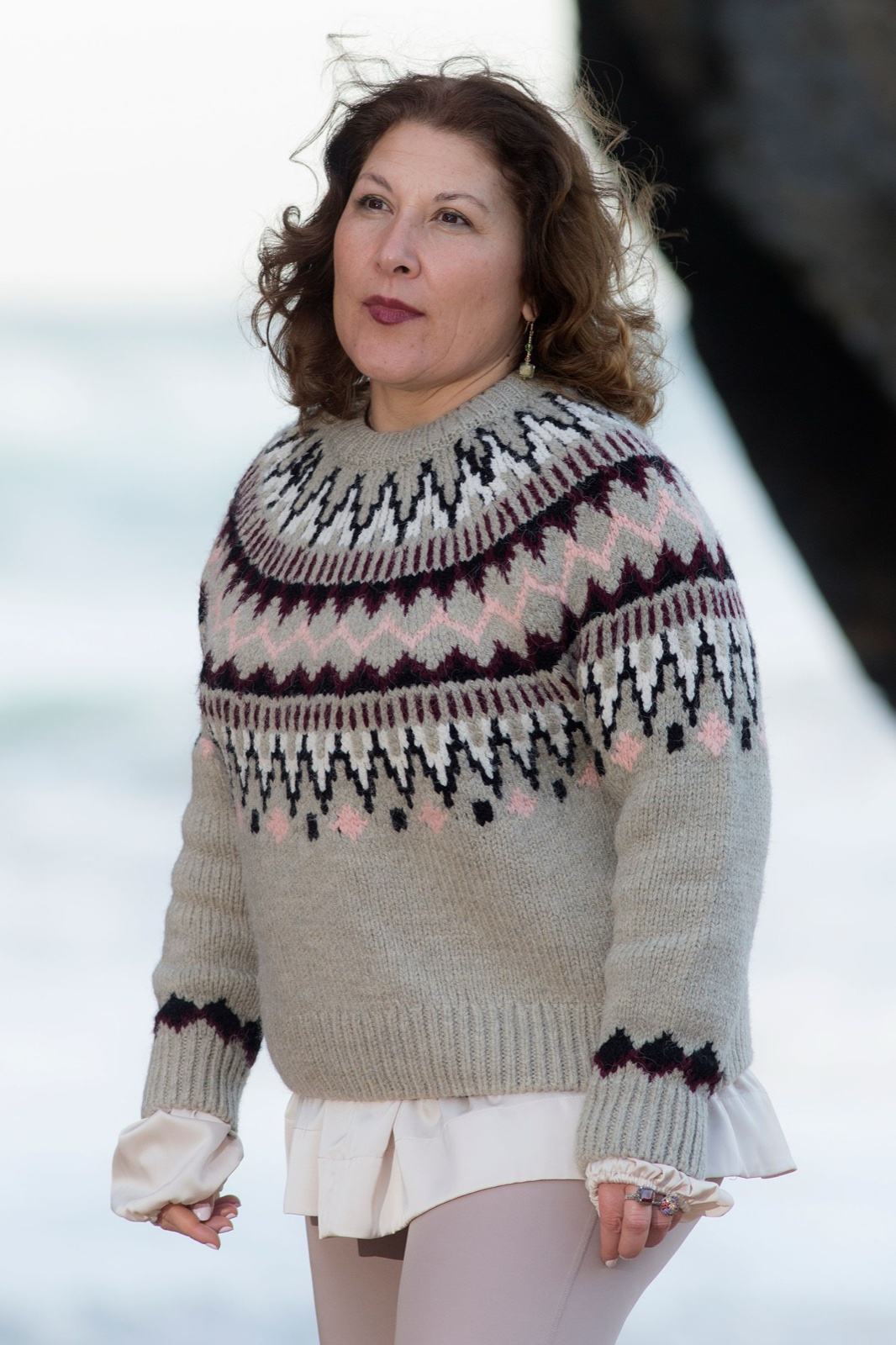 Woman standing on a beach in an oatmeal sweater with a multicolour stranded yoke in pink, burgundy, black and cream