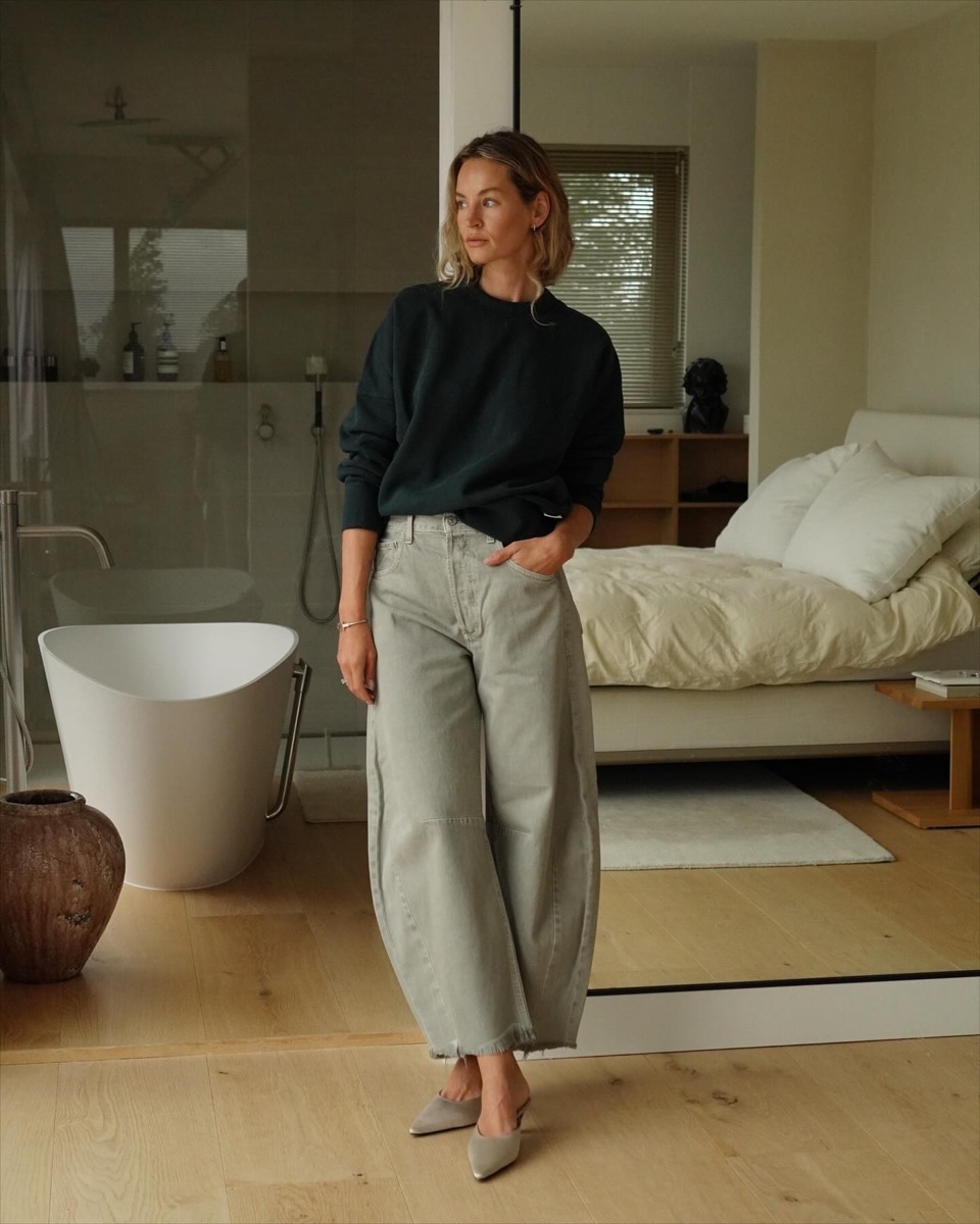 A woman in barrel-leg light-wash Citizens of Humanity jeans and a dark green sweatshirt standing in a bedroom in natural light